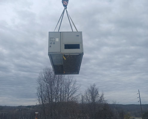 A crane lifts a large air conditioning unit above a rooftop, surrounded by cloudy skies and barren trees in the background.