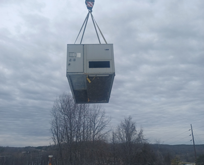 A crane lifts a large air conditioning unit above a rooftop, surrounded by cloudy skies and barren trees in the background.