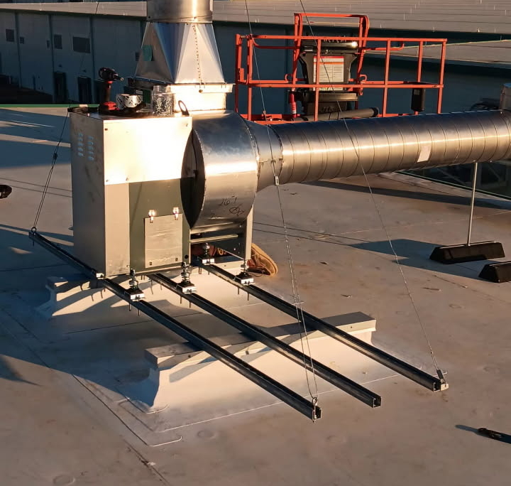 A metal ventilation unit with a tall chimney is mounted on a rooftop, with suspended support rods and a nearby lift in the background.