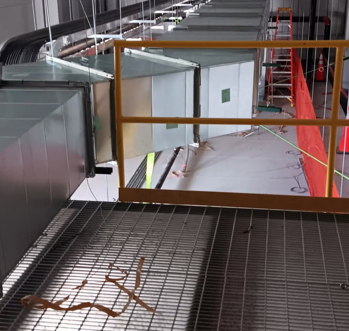 View from above showing suspended HVAC ductwork in a spacious industrial facility under construction, with ladders and safety barriers.