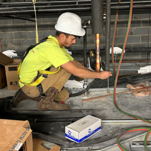 A worker in a bright yellow shirt is kneeling while adjusting pipes in a construction area, surrounded by boxes and tools.