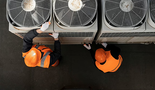 Two workers in safety gear inspect air conditioning units from above in a commercial setting.