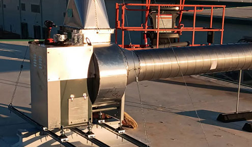 A metallic ventilation unit on a rooftop, featuring a large duct, with scaffolding and a backdrop of a building.