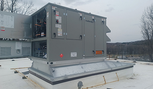 A large rooftop HVAC unit with an access panel, labeled "Valent," situated on a flat, white roof against a cloudy sky.