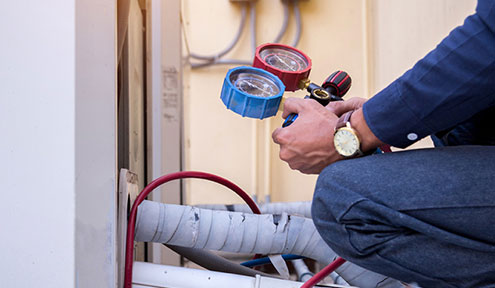 A technician checks refrigerant pressure using gauges, crouched beside an air conditioning unit with colored hoses.